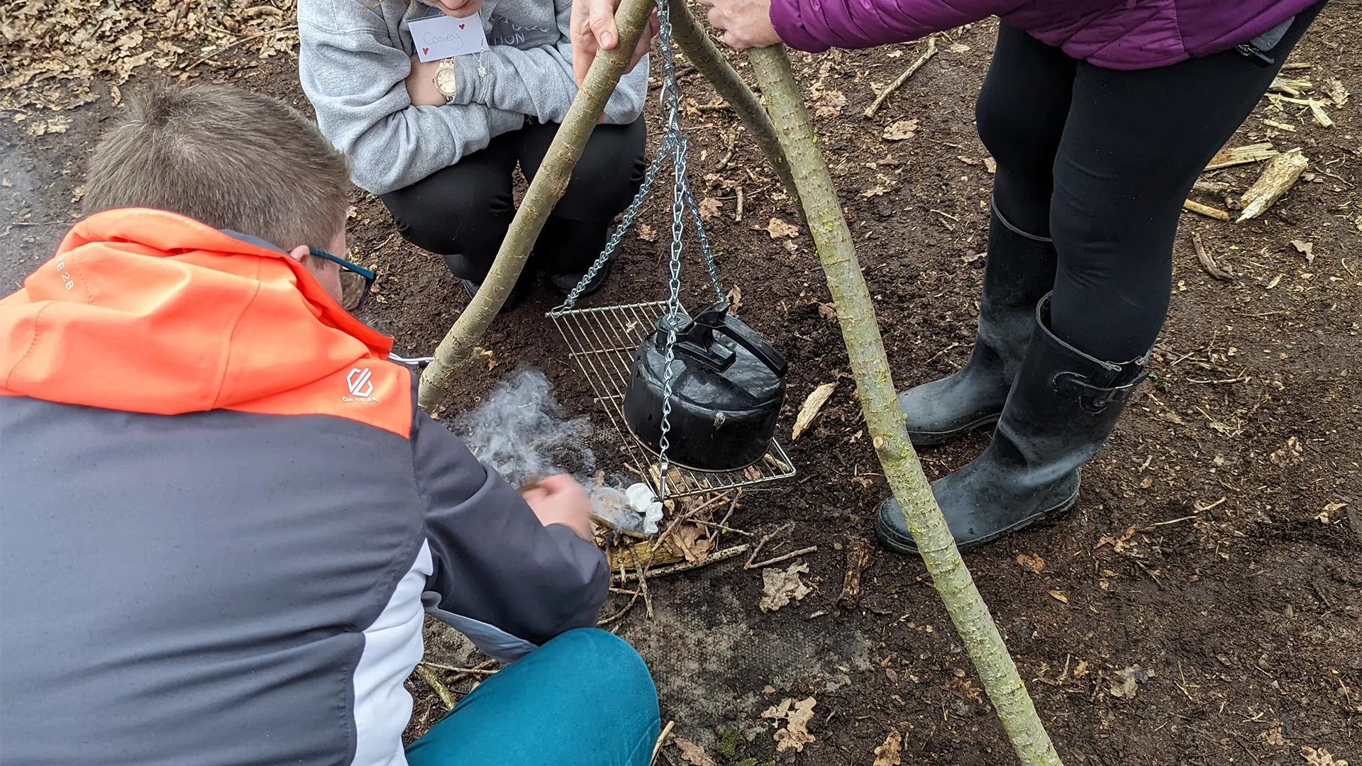 Three women light a fire outside under a camping kettle